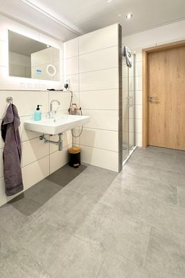 Das Bild zeigt ein modernes Badezimmer. Zu sehen ist ein Waschbecken mit einem Wasserhahn und einem Seifenspender darauf. Darüber hängt ein großer, beleuchteter Spiegel. Links neben dem Waschbecken ist ein Handtuch an einem Haken angebracht. Unter dem Waschbecken steht ein kleiner Mülleimer. Rechts befindet sich eine Duschkabine mit Glastür. Die Wände sind mit weißen Fliesen verkleidet, und der Boden besteht aus grauen Fliesen.