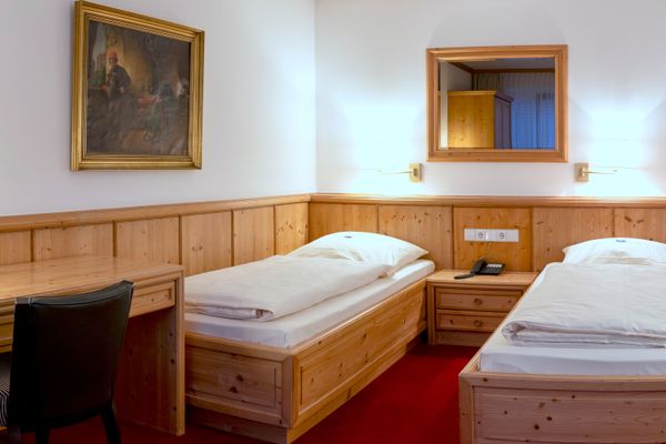 Das Bild zeigt ein Hotelzimmer mit zwei Einzelbetten, einem Nachttisch und einem Schreibtisch mit einem Stuhl. An den Wänden hängen ein großer Spiegel, Wandlampen und ein Gemälde. Der Boden ist mit einem roten Teppich ausgelegt.