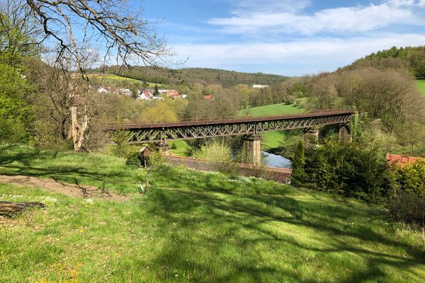 Eine alte, stählerne Eisenbahnbrücke führt über ein Tal mit einem Flusslauf. Die Landschaft drumherum ist geprägt von Wiesen, Feldern und Wald. Am linken Bildrand ist ein Ort erkennbar. Der Himmel strahlt blau.