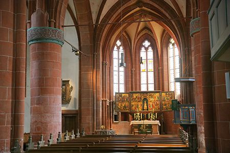 Vorbei an roten, hohen Säulen, über hölzerne Kirchenbänke hinweg, blickt der Betrachter in die Apsis der gotischen Kirche. Spitz zulaufende Sandsteinbögen treffen sich dort über drei hellen Fenstern und dem imposanten Flügelaltar, der sich über einem schlichten rechteckigen Sandsteinquarder befindet. Die große rechteckige, zentrale Tafel und die beiden halb so großen Seitenflügel sind üpppig mit bunten Personendarstellungen vor goldenem Hintergrund bemalt.
