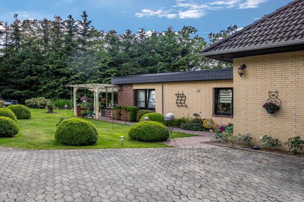 Gebäude mit gepflasterter Fläche im Vordergrund und gepflegtem Garten mit Sträuchern und Rasen.
