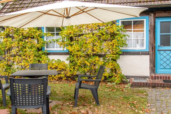Terrasse mit Tisch und Stühlen unter einem Sonnenschirm vor einer Hauswand mit Fenstern und Tür.