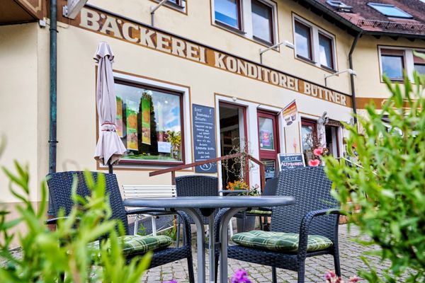 Blick durch Büsche auf Stühle und Bänke vor der Bäckerei.