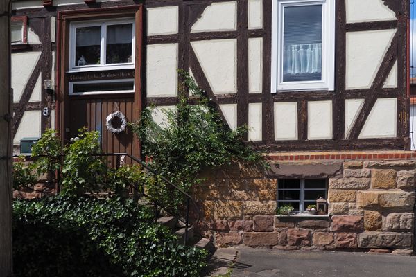 Ausschnitt eines braunen Fachwerkhauses mit Sandsteinsockel. eine einfache, mit Efeu berankte Treppe führt zur schlichten Eingangstür, die oberhalb ein geschnitzter Segensspruch ziert. Es steht geschrieben: Lebe und Liebe, für dieses Haus.