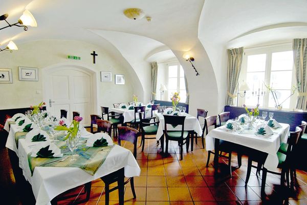 Ein Speisezimmer mit Gewölbedecke: Dunkle, festlich gedeckte Tische, Stühle mit grünen Sitzauflagen, rötlicher Fliesenboden und Sprossenfenster mit hellen Vorhängen.