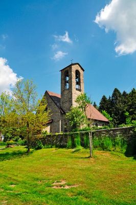 Im Vordergrund stehen einige junge Laubbäume in einer gemähten Wiese, dahinter umgibt die Friedhofsmauer die Filialkirche Rettenbach mit ihrem markanten Glockenturm.