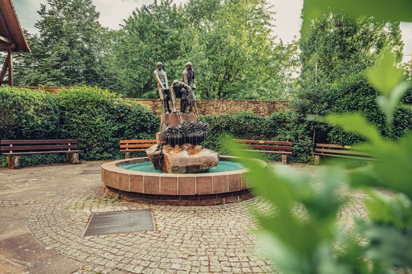 Ein Brunnen mit 3 Männern, um den Brunnen befinen sich Parkbänke