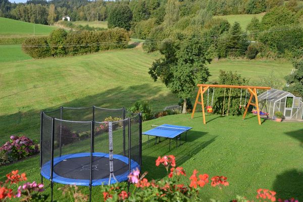 Von oben blickt man über Geranien auf eine Rasenfläche, auf der ein Trampolin, eine Tischtennisplatte, ein Holzschaukelgerüst mit drei Schaukeln und ein Gewächshaus stehen. Der Garten ist von Blumenbeeten und Sträuchern umgeben.