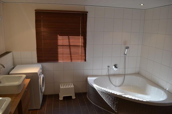 Badezimmer mit hellen Wand- und dunklen Bodenfliesen, Eckbadewanne, Waschmaschine, Doppelwaschbecken mit Unterschrank und holzfarbenem Innenrollo.