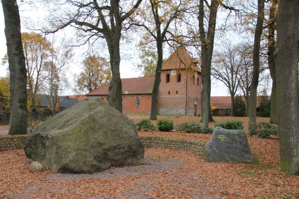 Großer Stein und kleiner Gedenkstein auf einer mit Herbstlaub bedeckten Fläche vor der Feldsteinkapelle St. Bartholomäus in Lüder.