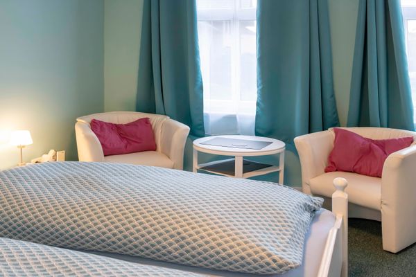 Helles Schlafzimmer mit türkisfarbenen Wänden. Im Vordergrund ein Bett mit blau gemusterter Bettwäsche. Dahinter stehen vor einem Fenster mit blauen Vorhängen zwei weiße Sessel mit rosa Kissen sowie ein runder, weißer Beistelltisch. Links leuchtet eine Nachttischlampe.