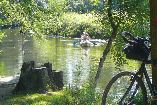 Ein Fahrrad lehnt an einem Baum am Ufer der Ilmenau, während zwei Paddler in einem Kanu vorbeifahren. Ein idyllischer Rastplatz mit Baumstümpfen und einem Seil zum Schwingen über das Wasser liegt im Schatten der Bäume.