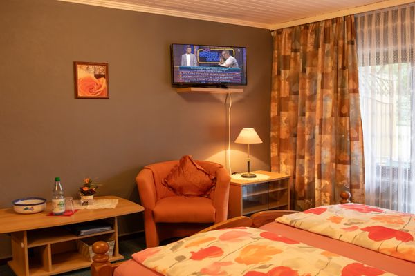 Schlafzimmer mit Bett, Sessel, kleinem Tisch und wandmontiertem Fernseher, mit Vorhängen und Innenbeleuchtung.
