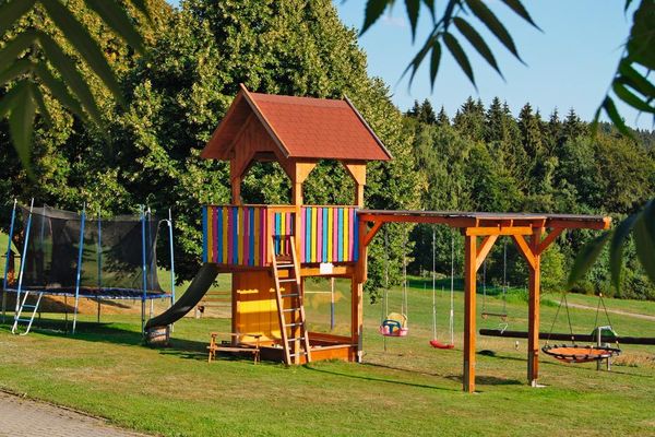Auf dem Rasenspielplatz, vor dem dichten Wald, stehen ein Trampolin, ein Spielhaus mit Rutsche und Schaukelgerüst, eine Vogelschaukel und eine Wippe.
