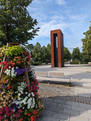 Das Bild zeigt den sogenannten Plessi-Turm, der aus Stahl besteht. Dieser steht am Eingang des ehemaligen Landesgartenschaugeländes in Kronach.. Man sieht im Vordergrund Blumen und Sitzbänke.