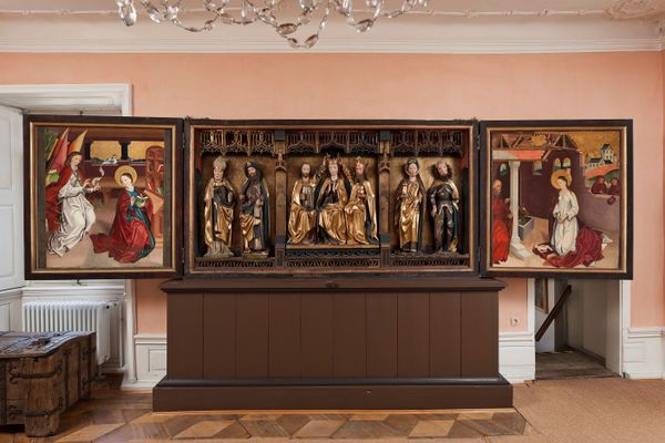Ein dreiflügeliger Marienaltar auf einem braunen Holzquader ist zentrales Bildmotiv. Die im Renaissance-Stil gemalten Bilder rechts und links sind sehr detailverliebt und erzählen jeweils eine Geschichte im Bild. Der zentrale Mittelteil besteht aus geschnitzten Figuren, mittig auf einem Thron mit Krone Maria. Der Altar steht in einem rosa gehalteten Raum mit Holzboden und weißen Stuckelementen. Links neben dem Altar ist eine schwere hölzerne Truhe aus dem Mittelalter mit Metallverzierungen, dahinter ein Fenster. Rechts des Altares befindet sich eine Tür mit einem Treppenabgang.