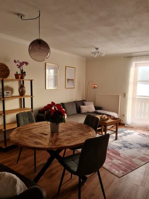 In Raum befinden sich ein graues Sofa, eine Stehlampe und ein kleiner Holztisch. Daneben steht runder Esstisch mit drei Stühlen. Ein fünfstöckiges Holzregal mit Dekoartikeln steht an der Wand, und auf dem Fußboden liegt ein rot-schwarz gemusterter Teppich. Über dem Sofa hängen zwei Bilder. An der Decke befinden sich eine runde Lampe und ein Strahler.