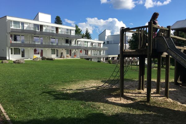 Ein Spielplatz auf einer Rasenfläche vor einer Apartmentanlage. Ein Kind steht auf einem Holzkletterpodest am Einstieg einer Rutsche, bereit zum Herunterrutschen.