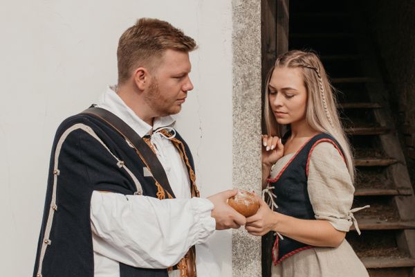 Ein mittelalterlich gekleideter Mann reicht einer traurig dreinblickenden Frau ein Stück Brot. Sie lehnt still an einer steinernen Türöffnung neben ihm.