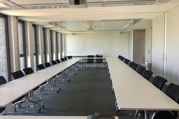 Großer, heller Konferenzraum mit zwei langen Tischen in U-Form und vielen schwarzen Stühlen.