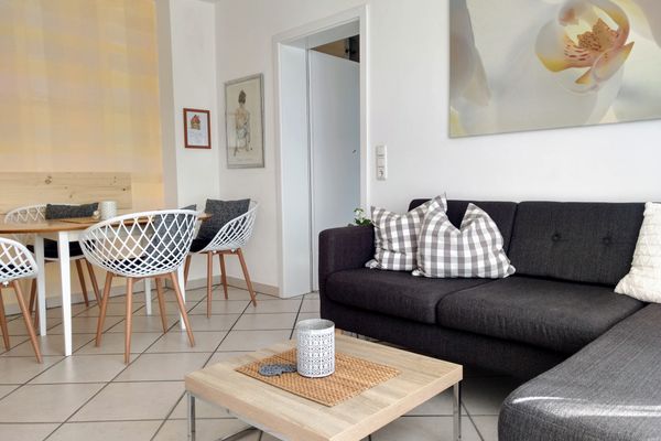 Ein graues Ecksofa, davor ein viereckiger Holztisch mit Edelstahlgestell sowie ein Esstisch und Stühle in Weiß und Holz stehen in einem Raum mit hellen Bodenfliesen. Über dem Sofa hängt ein Bild mit Orchideenmotiv. Zwei kleinere Bilder schmücken eine Wand über dem Essbereich.