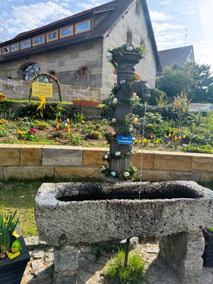Steinerner Trog mit metallener Säule und Drachenkopf-Wasserspeier. Säule umwickelt mit einer Buchsbaumgirlande mit bunten Ostereiern.