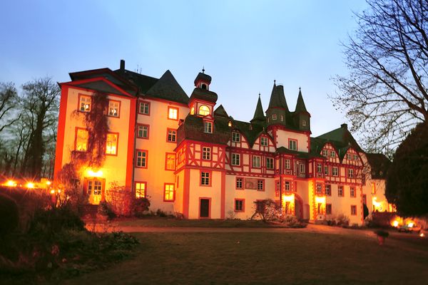 Hinter winterlich kahlen Bäumen ist ein mit warmen Licht angestrahlter Schlosskomplex bei Nacht zu sehen. Die Gebäude sind aus massivem Stein gebaut und haben dunkelgraue Schieferdächer. Die verschachtelten und in den Höhen abgesetzten Gebäudeteile sind weiß verputzt. Alle Fensterleibungen und Kanten sind aus rotem Sandstein. Gerade in den mittleren Teilen sind Obergeschosse und Dachgeschosse mit rotem Fachwerk aufesetzt. Einige Türme und Zinnen sind besonders im mittleren Teil präsent, während die Seitenteile zwar mächtiger, aber schlichter sind.