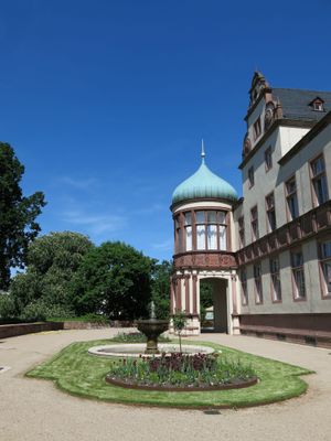 Teilansicht eines historischen Schlossgebäudes mit rundem Erker, blauem Kuppeldach, Springbrunnen und Blumenbeet im Innenhof bei blauem Himmel