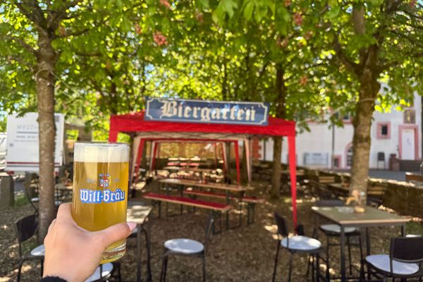 Auf dem Foto ist der Biergarten im Hintergrund abgebilder. Im Vordergrund wird ein Glas Bier hochgehalten
