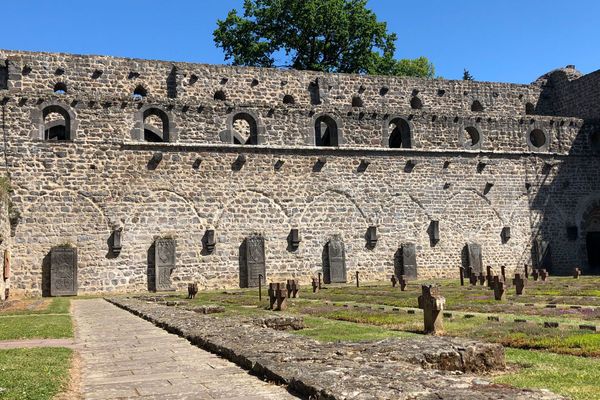 Die Ruinen der Klosterkirche. Eine große Wand mit vielen Fenstern und ein paar Grabplatten davor ist zu sehen. Auf einem Rasenstück mit gepflasterten Gehwegen davor stehen vielen Steinkreuze in Reihen. Es ist wohl ein Friedhof