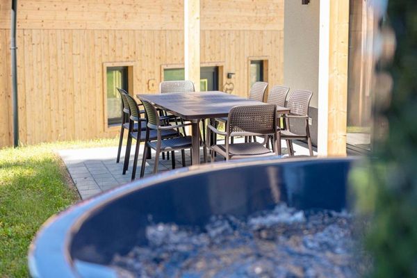 Im Vordergrund ist ein Teilausschnitt eines grauen Hot Pot zu sehen. Im Hintergrund ein großer brauner Terrassentisch mit acht Stühlen, Ausblick auf das Nachbarchalet.