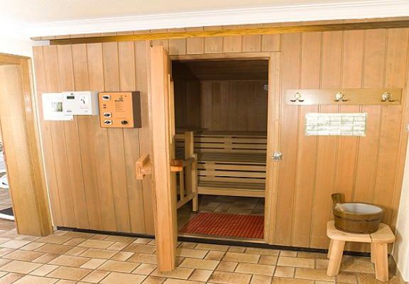 Die Tür zur Sauna steht offen, daneben befindet sich auf einem Holzhocker ein kleiner, runder Holzbottich mit Griff.
