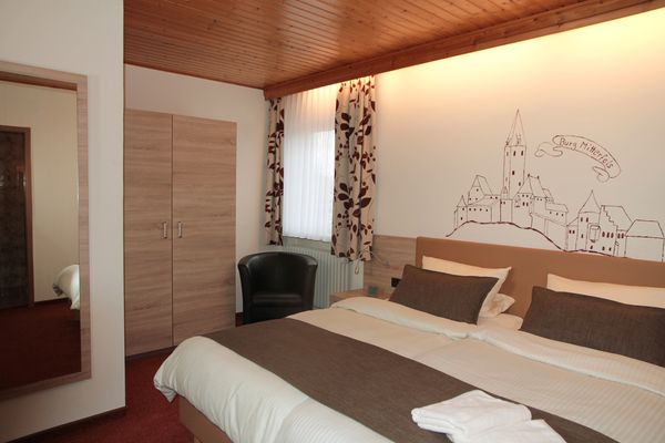 Auf dem hell bezogenen Doppelbett mit braunen Zusatzkissen liegen zwei Handtücher. Über dem Bett hängt eine Skizze der Burg Mitterfels. Ein schwarzer Sessel steht vor einem hellen Holzschrank und einem Fenster mit gemusterten Vorhängen. Ein Ganzkörperspiegel ergänzt die Einrichtung.