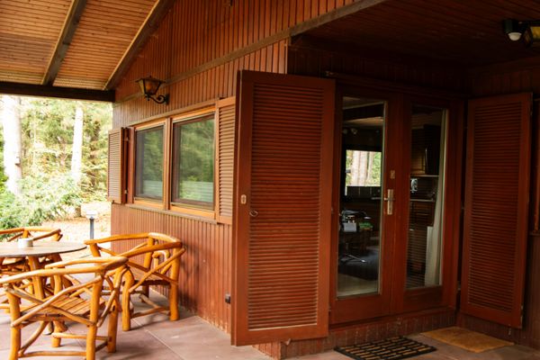 Überdachte Terrasse mit Holztisch und Stühlen vor einer Holzfassade mit Tür und Fensterläden.