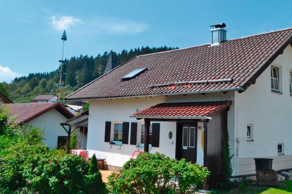 Ein Einfamilienhaus mit Sprossenfenstern und teilweise Fensterläden. Der Eingangsbereich und die Terrasse an der Stirnseite sind überdacht. Im Vordergrund wachsen üppige Sträucher. Im Hintergrund sieht man ein Spielhaus mit Rutsche und neben weiteren Gebäuden einen Maibaum und einen Kirchturm.