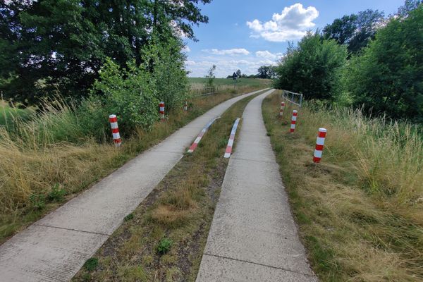 Zweispuriger Beton-Wirtschaftsweg mit Grasstreifen in der Mitte, flankiert von roten Markierungspfosten und niedrigen Metallsperren, links und rechts hohes Gras und Büsche, der Weg führt in eine sanfte Kurve durch offene Feldlandschaft unter blauem Himmel mit Wolken.