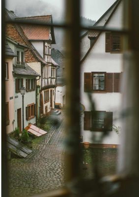 Das Bild zeigt eine ruhige, enge Gasse mit Fachwerkhäusern, gesehen durch ein Fenster. Die Stimmung wirkt ruhig und leicht verträumt.