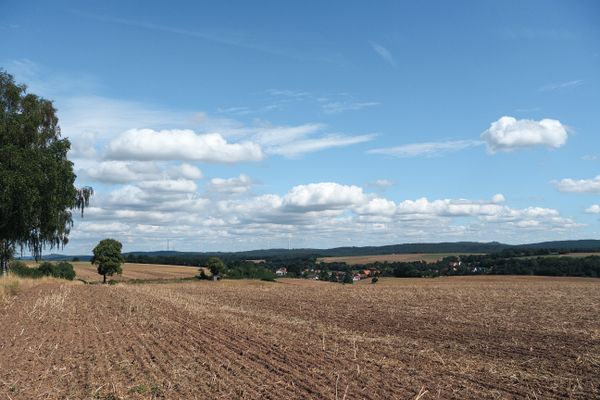 Ein weiter Blick in die Landschaft, die maßgeblich aus Stoppelfeldern besteht. Hier und da stehen zwischen den Äckern ein paar Bäume und büsche, in der Ferne liegt ein Dorf. Der Himmel ist herrlich blau mit Gutwetterwolken.