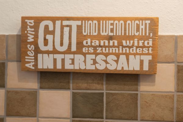 Holzschild mit weißem Schriftzug an einer Wand mit mehrfarbigen Fliesen im Hintergrund.