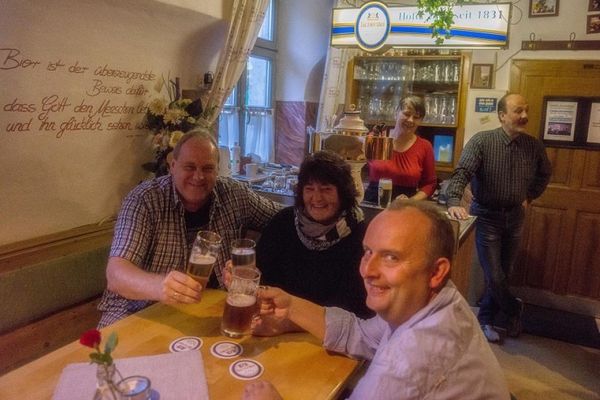 Auf dem Bild sieht man im vorderen Bereich 3 Gäste an einem Tisch sitzend, die mit Blick zum Fotografen, mit ihren Getränken (Bier) anstoßen. Im Hintergrund ist der Wirt zu sehen. Daneben steht der Tresen mit der Zapfanlage, an der eine Frau ein Bier zapft.