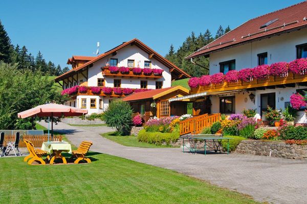 Zwei große, nebeneinander liegende Gebäude mit rotblühenden Blumen auf den Balkonen. Vor den Häusern befinden sich schön gepflegte, bunt bepflanzte Beete, davor eine Tischtennisplatte. Zwischen den Häusern steht ein Gartenhaus. Gegenüber befindet sich eine Liegeweise mit Sitzgelegenheiten unter Sonnenschirmen.
