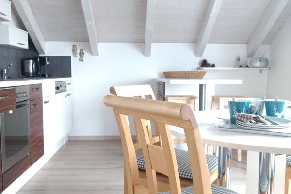 Unter einer Mansarde mit heller Holzdecke und Sichtbalken befindet sich eine weiße Küchenzeile, bei der einige der unteren Schranktüren aus dunklem Holz sind. Gegenüber steht eine Esstheke, davor ein runder, weißer Tisch mit Holzstühlen. Dekoartikel und ein CD-Player stehen auf einem Wandbord.