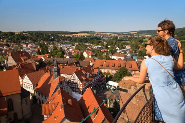 Ein Paar blickt von einem Turm über die vielen roten Dächer des Fachwerkortes weit ins Umland. Viele Felder, teils schon Gelb, und Wälder umgeben die mittelalterliche Kleinstadt Schlitz.
