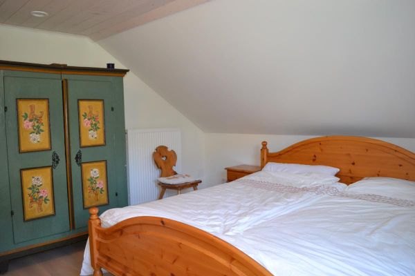 Unter einer Dachschräge steht ein Kieferholz-Doppelbett mit Nachtkästchen und Stuhl; daneben ein bemalter Bauernschrank.