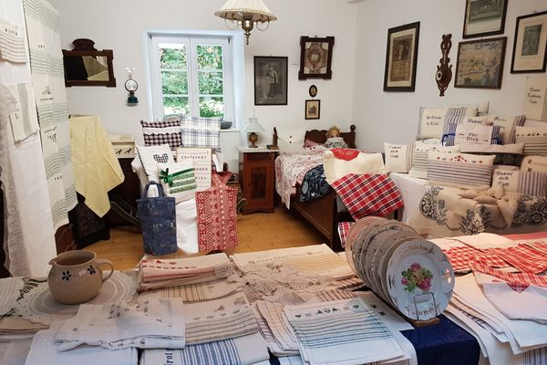 Zu sehen ist ein historisch eingerichteter Raum mit alten Bildern an der rechten Wand, einem alten Holzbett darunter und links daneben einem Nachttisch mit Leuchte. Überall im Raum sind unterschiedlichste Tücher mit und ohne Spitze, Karo oder Lochmuster zu sehen. Auch alte Teller mit Malerei und ein irdener Topf werden auf einem über und über mit Tüchern belegten Tisch im Vordergrund präsentiert.
