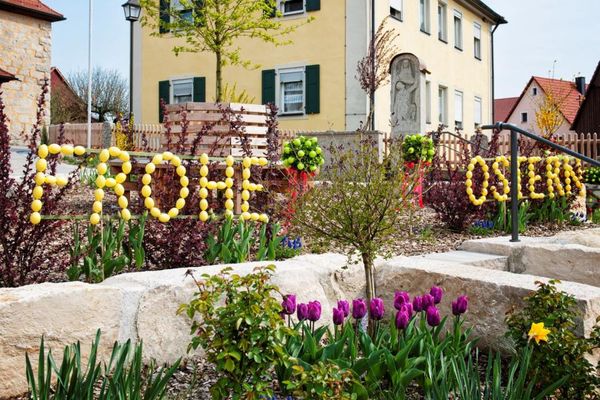 In einem Blumenbeet ist in großen Buchstaben aus vielen gelben Eiern der Schriftzug "Frohe Ostern" angebracht.