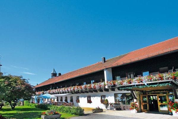 Außenansicht des langgezogenen Hotelgebäudes mit blumengeschmücktem Eingangsbereich, Terrasse und herrlich blühenden Blumen auf den Balkonen. Die gepflegte Rasenfläche wird von Bäumen und Sträuchern beschattet.