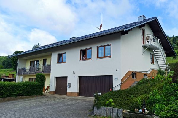 Dreigeschossiges Haus mit Hecke, Terrasse und Balkon. Neben der Terrasse zwei Garagen. Eine Treppe führt zum Obergeschoss, eine Außen-Edelstahltreppe zum Dachgeschoss. Rechts neben dem Gebäude ein Brunnen mit Steintrog in einer bepflanzten Böschung.