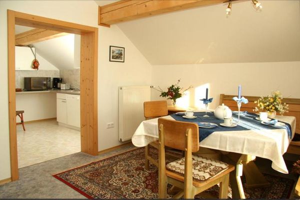 Mansarden-Esszimmer mit einer Holz-Eckbank, einem Tisch und zwei Stühlen. Ein großer Durchgang führt in eine Küche mit weißen Schränken.