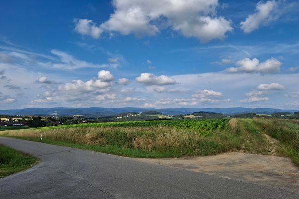 Eine Kreuzung führt in ein Feld und auf eine schmale Straße, die zu verstreuten Ortschaften zwischen Wiesen und Waldstücken führt. Am Horizont erstreckt sich eine sanfte, bewaldete Hügellandschaft.